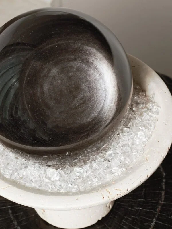 Stone Blessings close up of a polished obsidian sphere resting on clear quartz chips in a travertine pedestal bowl